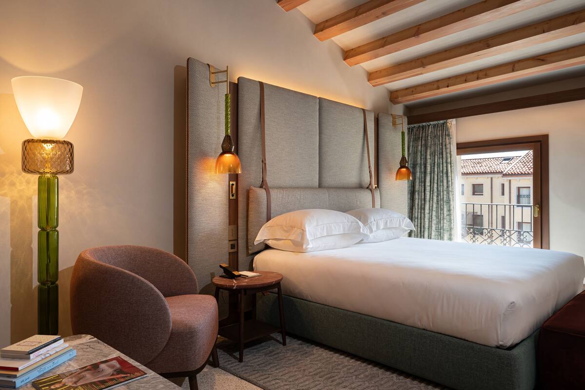 A hotel room with a large bed, two bedside lamps, a chair, a small table with books, and a view of buildings through a window.