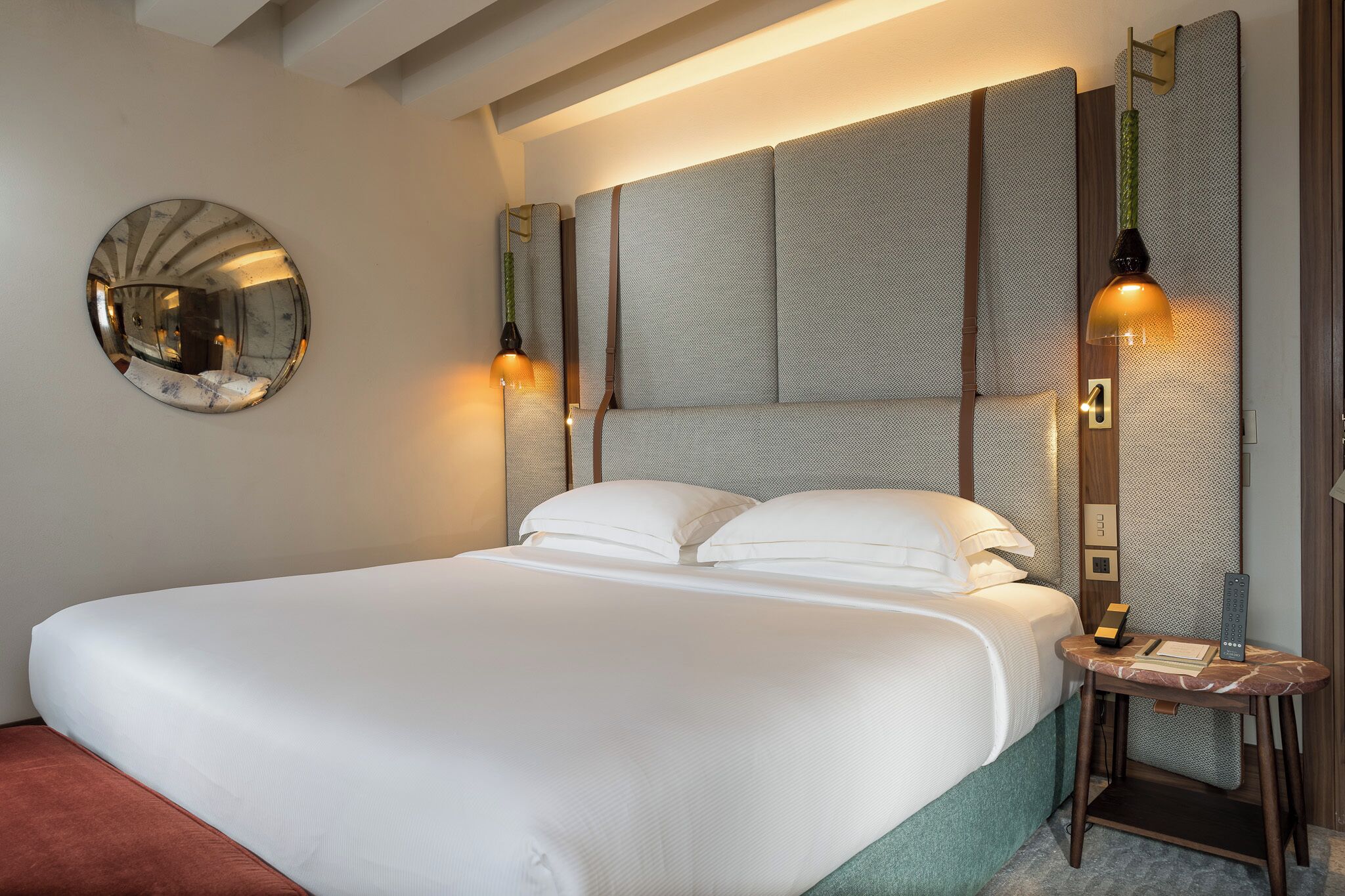 A neatly made bed with white linens, a round mirror on the wall, and a bedside table with a lamp and a book.