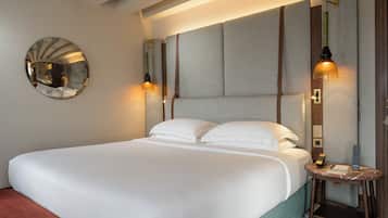 A neatly made bed with white linens, a round mirror on the wall, and a bedside table with a lamp and a book.