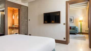 Habitación de hotel con una cama grande, un televisor de pantalla plana en la pared, un baño con tocador y perchero para toallas, y una sala de estar con sofá y lámpara.