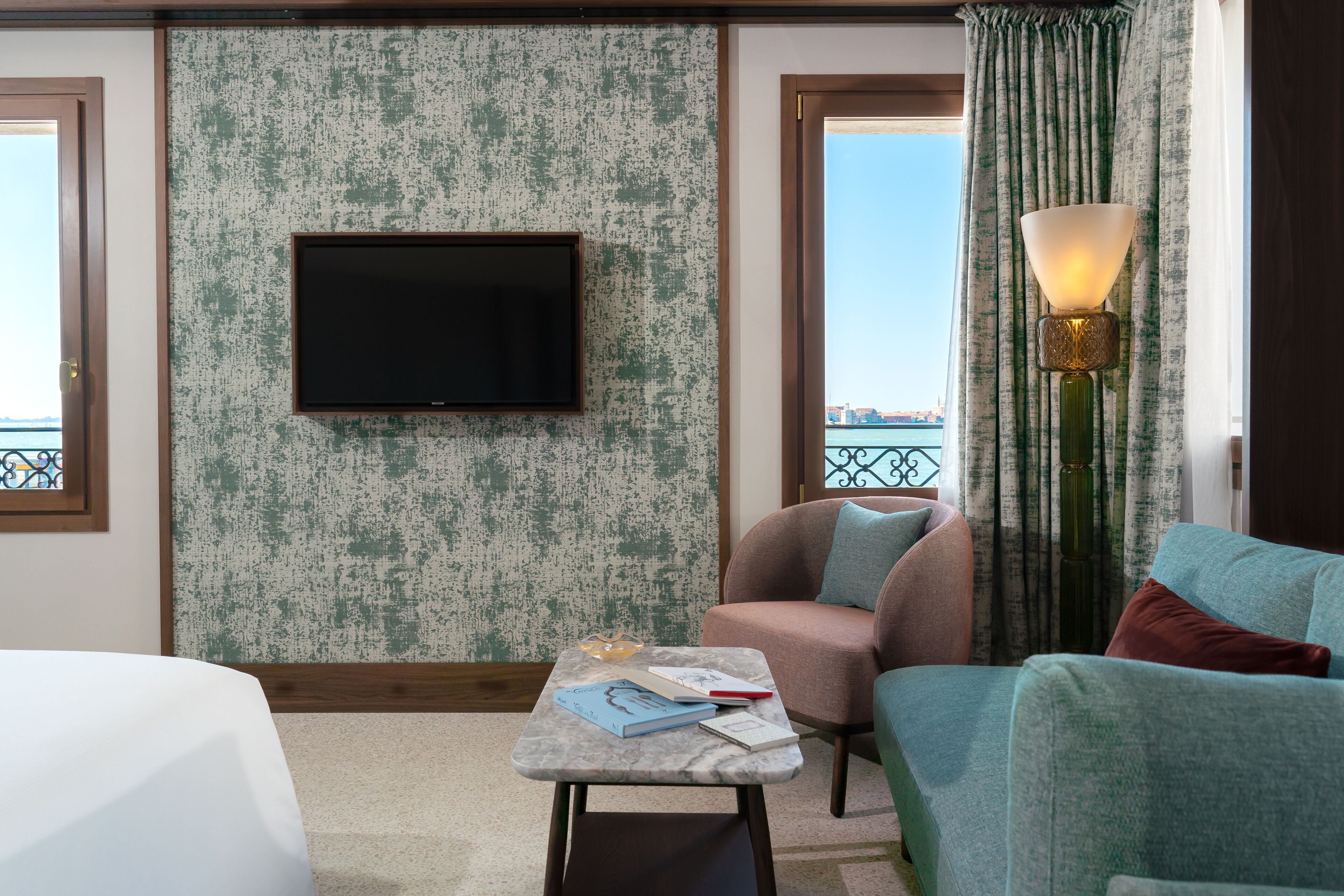 Quarto de hotel com uma televisão grande, uma mesinha com livros, um sofá e um abajur.