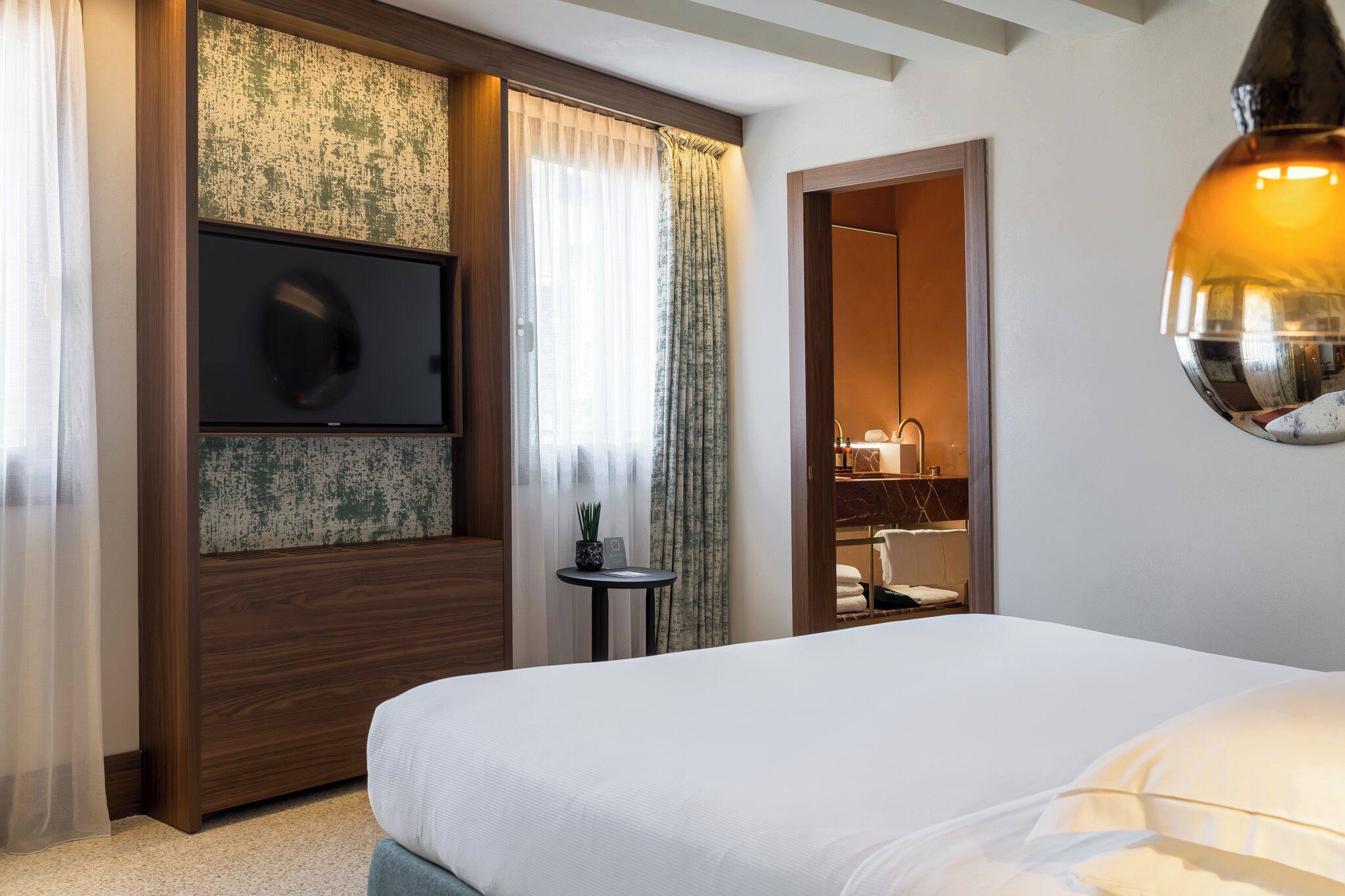 Une chambre d’hôtel avec un grand lit, un téléviseur fixé au mur, une petite table avec une plante et un miroir dans un coin.