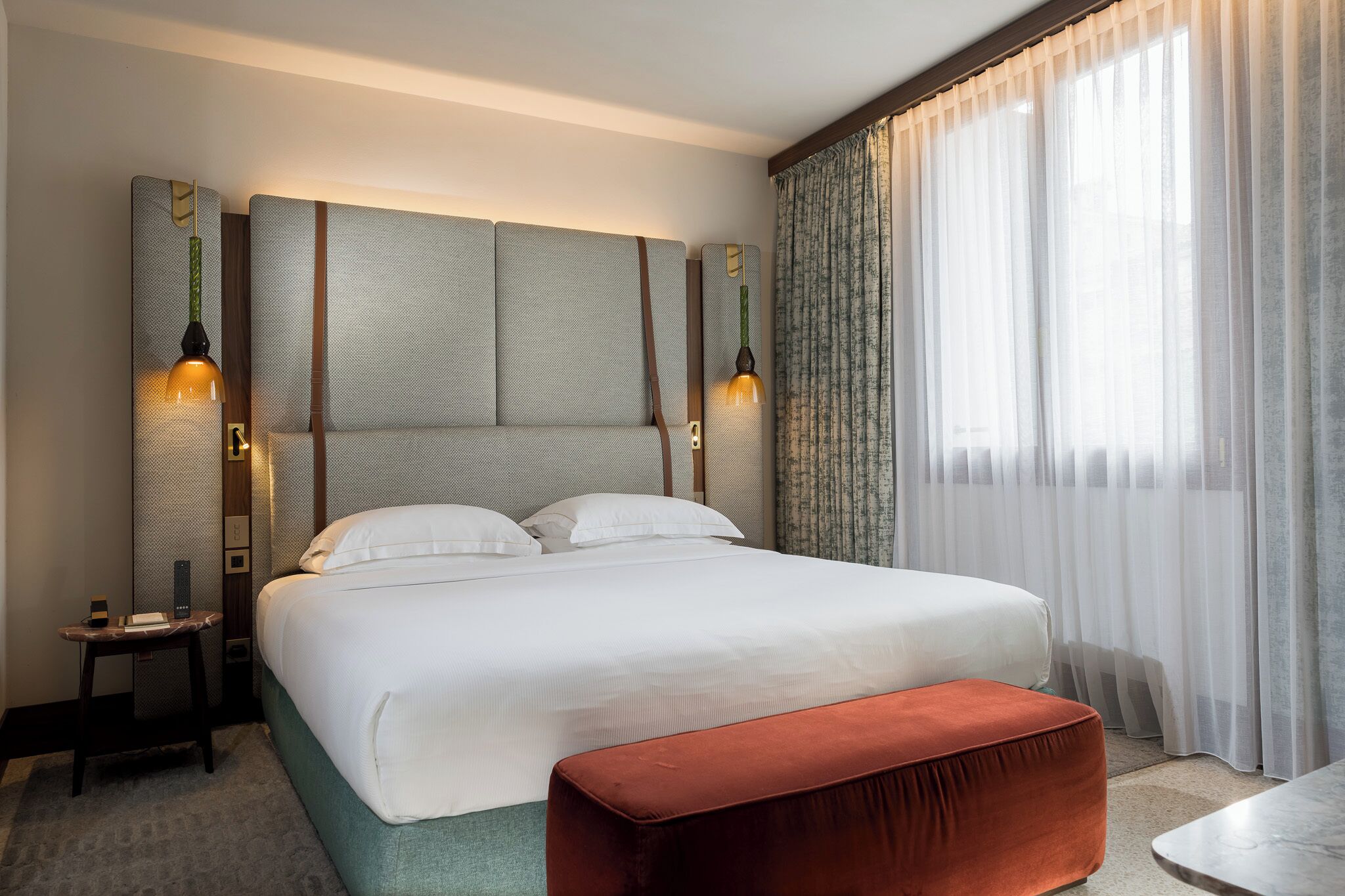 Habitación de hotel con una cama grande, una mesita de noche, una lámpara de lectura, un banco y una ventana con cortinas translúcidas.