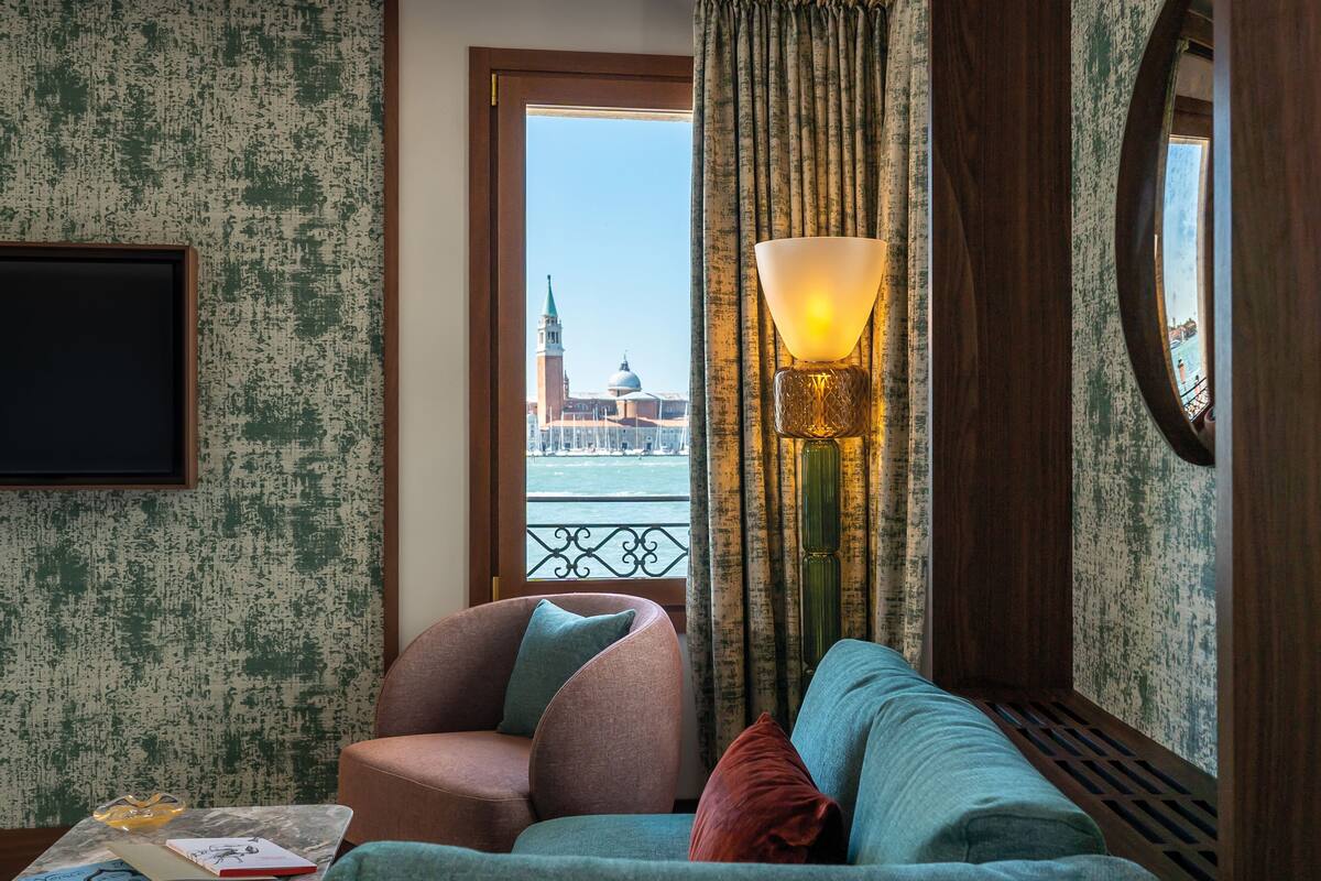 A room with a view of a historic building, a sofa, a chair, a lamp, and a small table with a book and glasses.