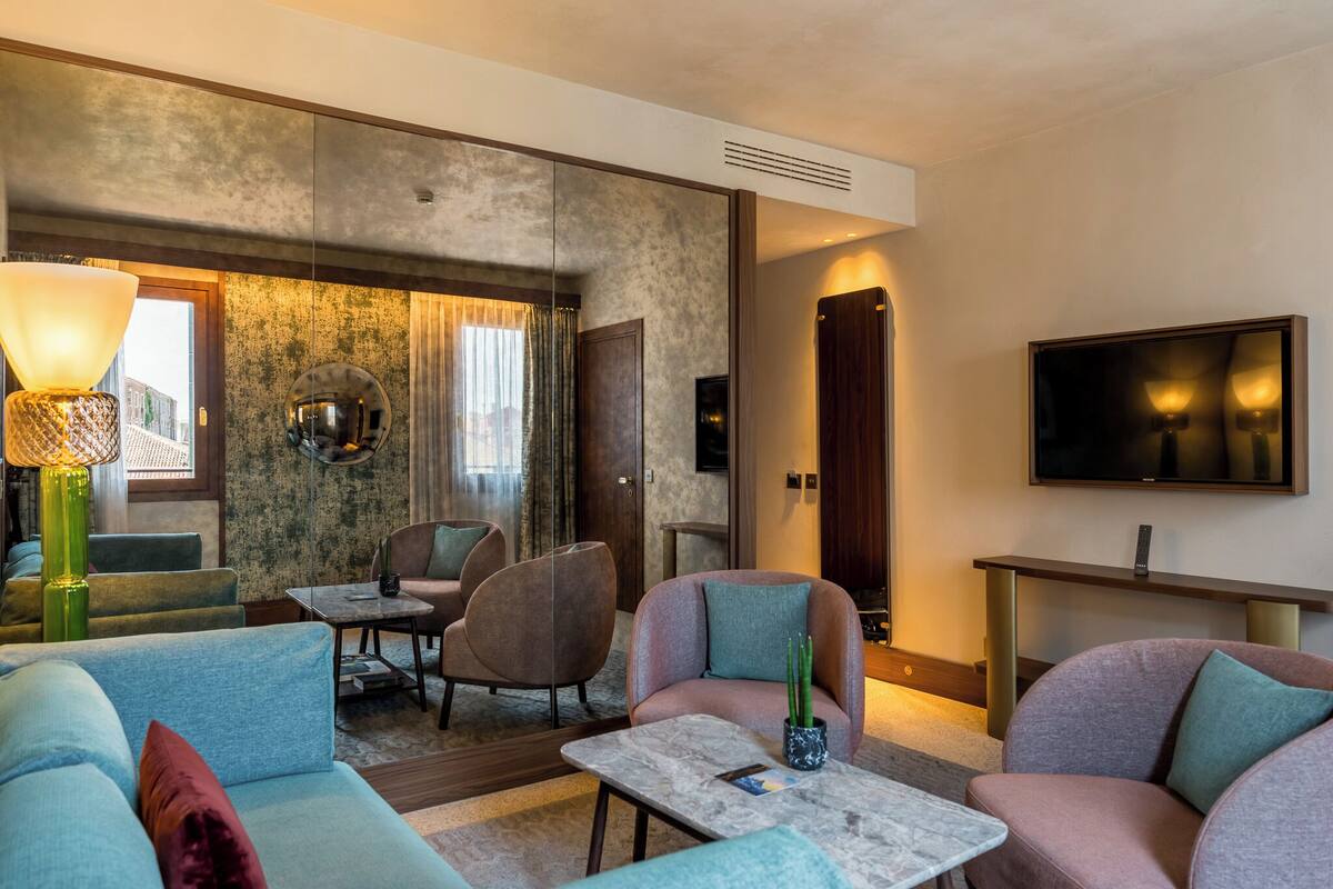 A modern hotel room with a sofa, armchairs, a coffee table, a TV, and a large window with curtains.