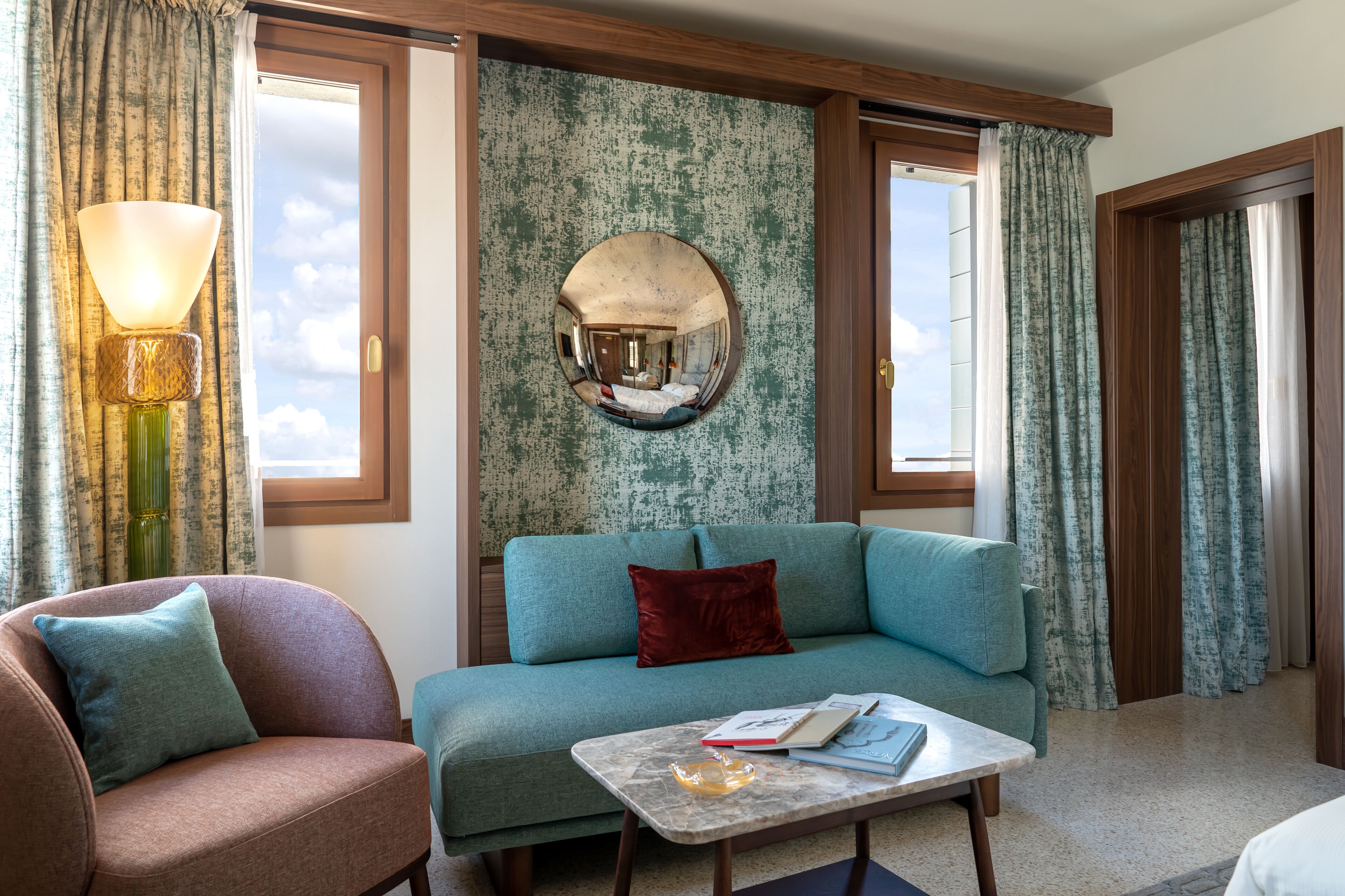 A room with a teal sofa, a brown armchair, a round mirror, and a table with books.
