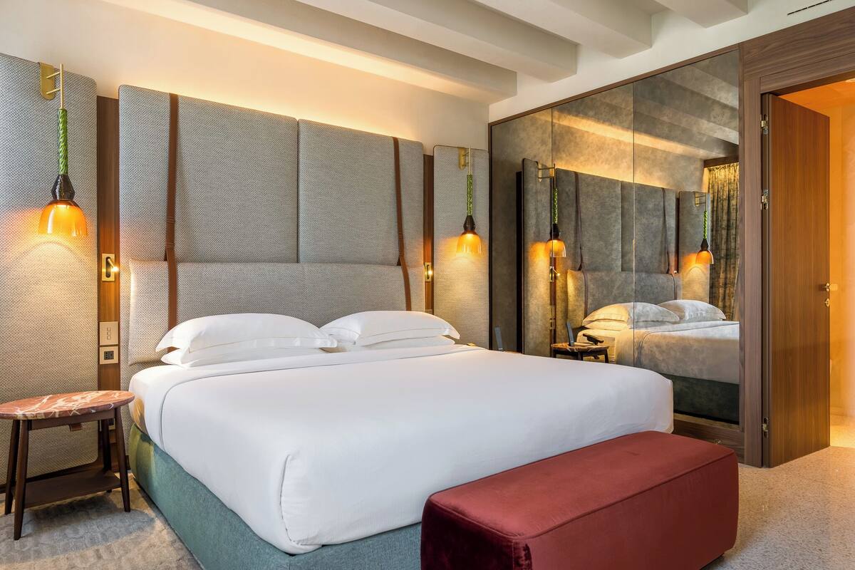 A modern hotel room with a large bed, a red bench, a small table, and a mirror reflecting the room.