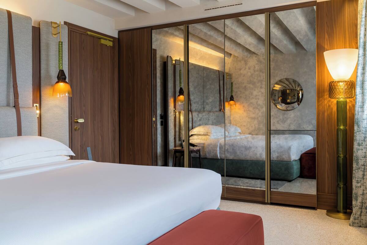 A hotel room with a large bed, a wardrobe, a mirror, and a nightstand with a lamp.