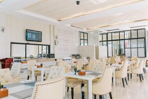 A spacious dining area with multiple tables and chairs, a flat-screen TV mounted on the wall, and large windows providing natural light. - Aramis Hotel (Nakhon Sawan)