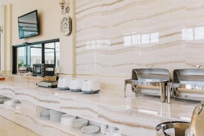 A hotel breakfast area with a marble countertop, buffet setup, and a television mounted on the wall. - Aramis Hotel (Nakhon Sawan)