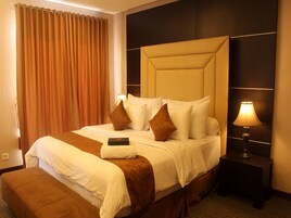 Una habitación de hotel con una cama grande, una mesita de noche con lámpara, un banco al pie de la cama y una cortina.