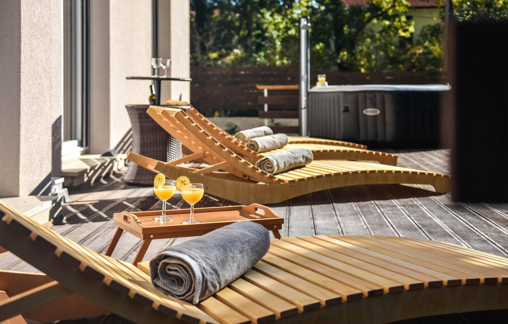 A sunlit outdoor lounge area with wooden loungers, a small table with glasses of orange juice, and a barbecue grill in the background.