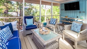 A covered outdoor seating area with wicker furniture, a glass coffee table, and a television mounted on the wall. - Family-Friendly; Steps to Beach; Fabulous Backyard w/ Screened Porch, Expansive Back Deck & Fire Pit (Isle of Palms)
