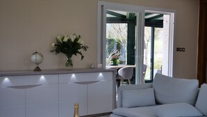 A modern living room with a white sofa, a glass coffee table, and a small kitchen area with a counter and a vase of flowers. - MIL ROSES EN BAIE 9 PEOPLE NEAR MARQUENTERRE (SAINT QUENTIN EN TOURMONT)