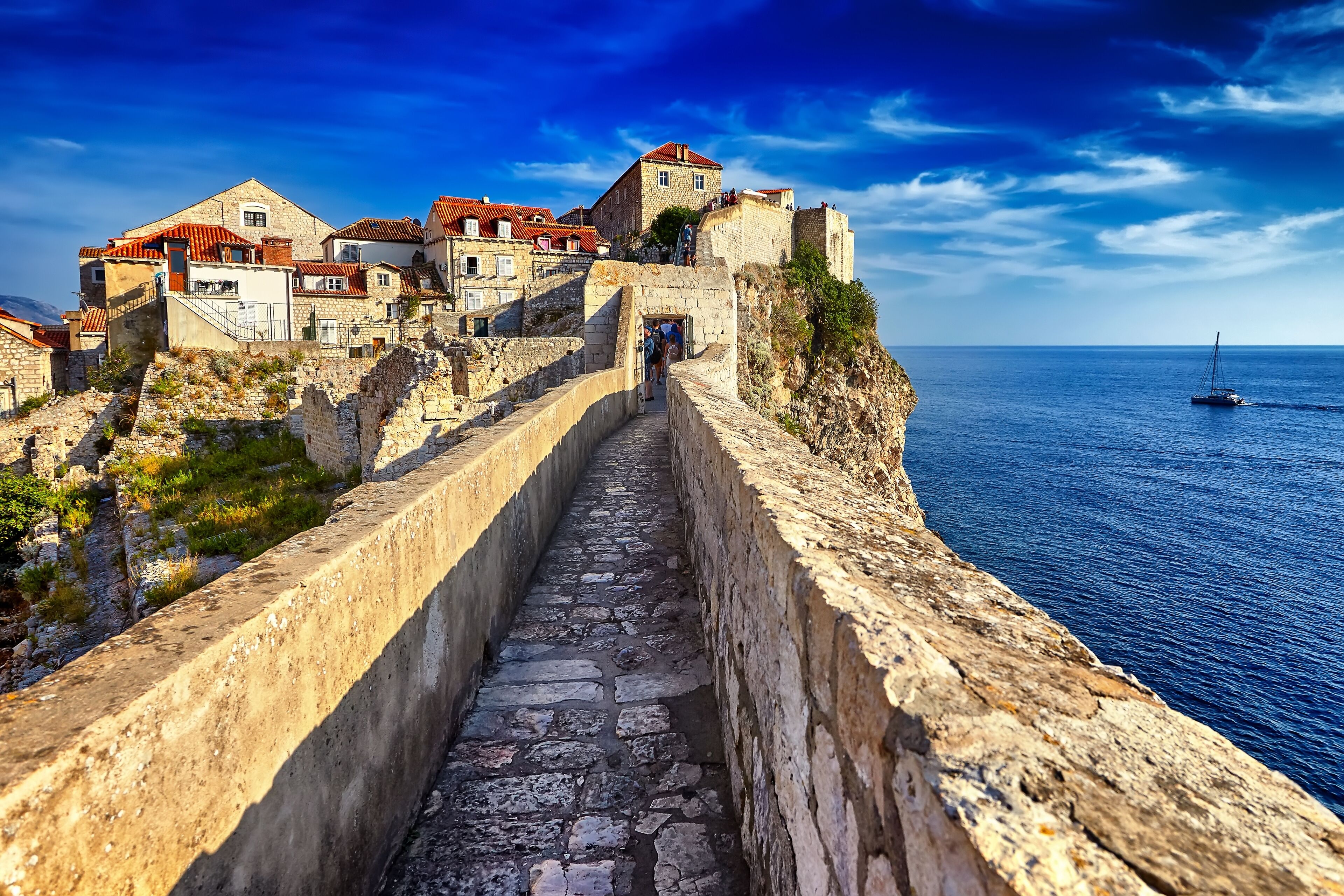A stone pathway leading to a historic building complex with red-tiled roofs, overlooking a calm sea.