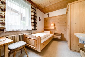 A wooden room with a bed, a desk, a chair, a window with curtains, a wall-mounted cross, and a wall-mounted light fixture.