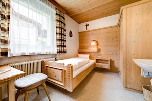 Una habitación de madera con una cama, un escritorio, una silla, un radiador, una lámpara fijada a la pared y un armario de madera.