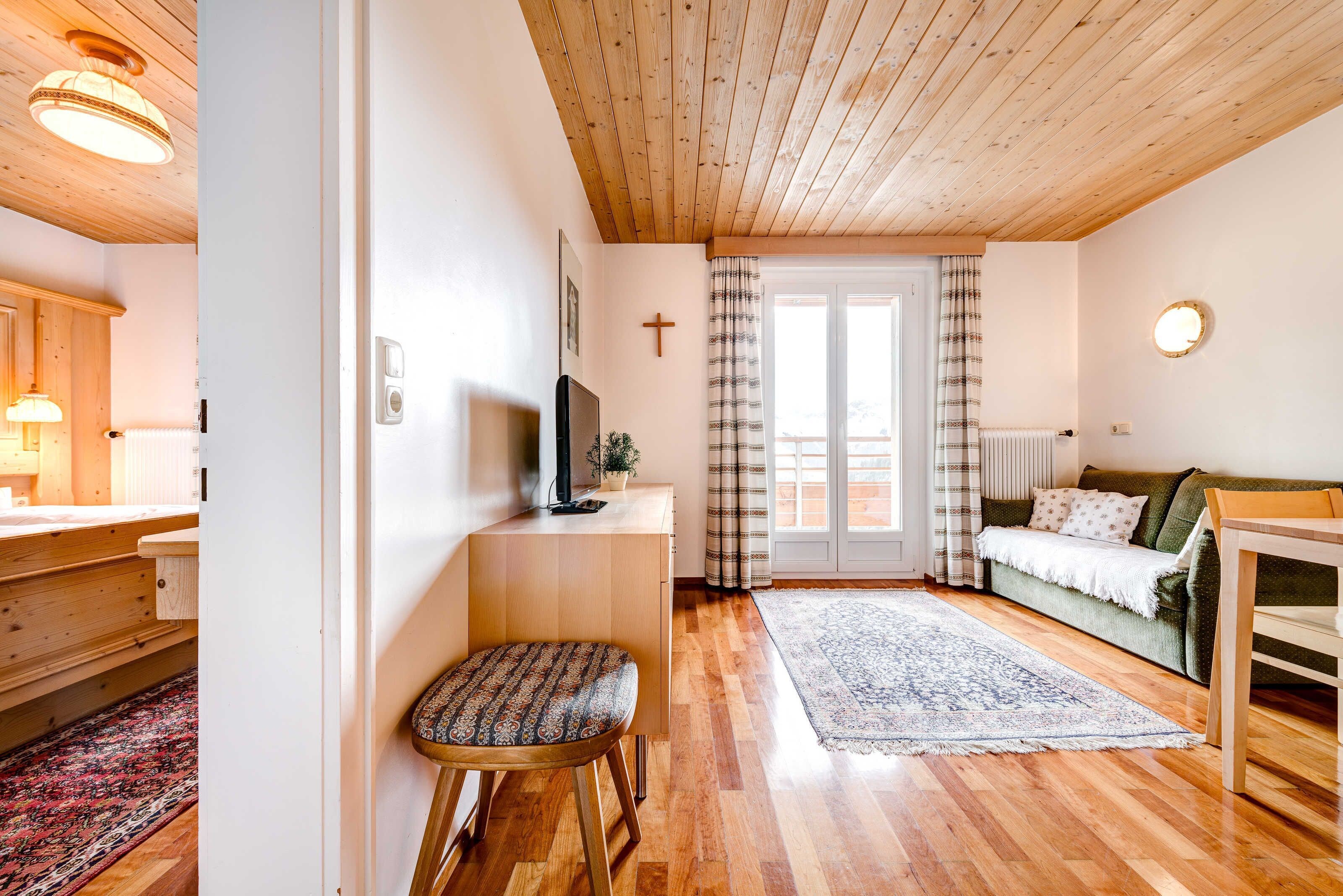 Una habitación con suelo de madera, un sofá, una mesita, un televisor, una cruz en la pared y una ventana con cortinas.