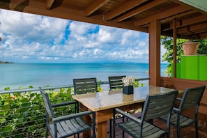 A covered outdoor seating area with a table and chairs, overlooking a body of water and a distant landmass. - Villa Dos Palmas - Beach  Villa - Waves Lapping at Your Door (Vieques)