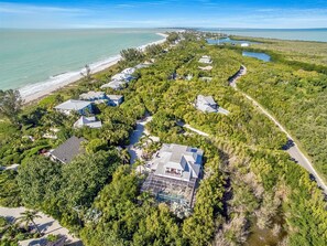An aerial view of a coastal residential area with houses surrounded by dense greenery, a beach, and a clear view of the ocean.