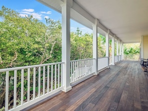 A covered porch with white railings and wooden flooring, surrounded by lush greenery.