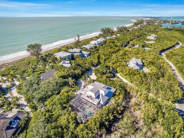An aerial view of a coastal resort with multiple buildings surrounded by dense greenery, a beach, and a clear view of the ocean.