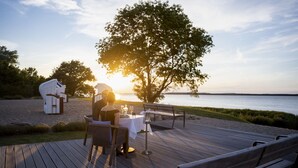 Une terrasse en bois avec une table dressée pour deux personnes, donnant sur une étendue d’eau et un arbre.