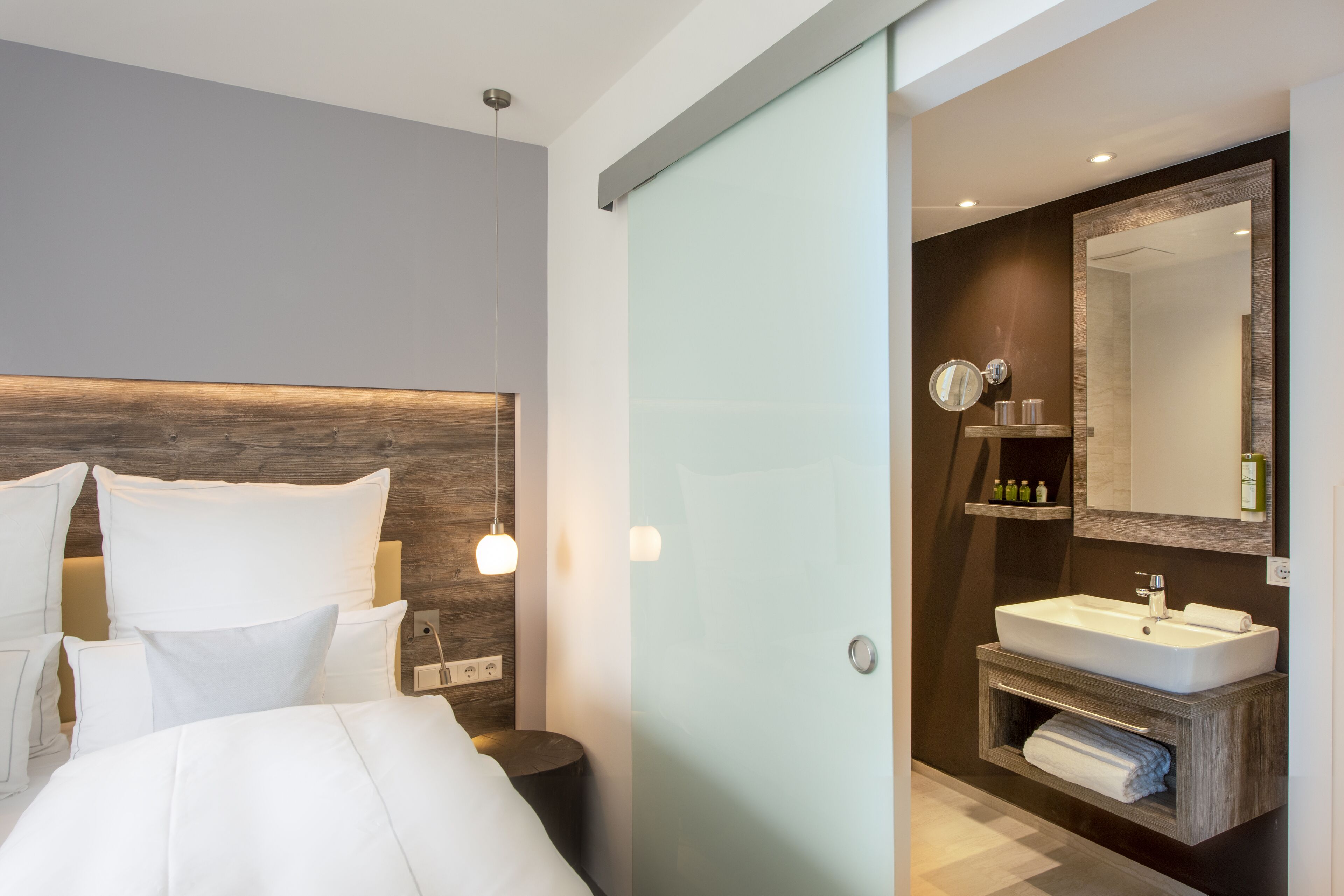 Un dormitorio moderno con una cama grande, lámparas de noche y una mesita de noche. Junto al dormitorio, un baño con un espejo grande, lavamanos y un perchero para toallas.