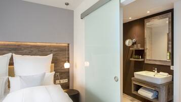 Un dormitorio moderno con una cama grande, lámparas de noche y una mesita de noche. Junto al dormitorio, un baño con un espejo grande, lavamanos y un perchero para toallas.