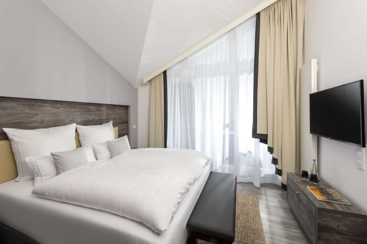 A modern hotel room with a large bed, a flat-screen TV, a wooden headboard, and a large window with sheer curtains.
