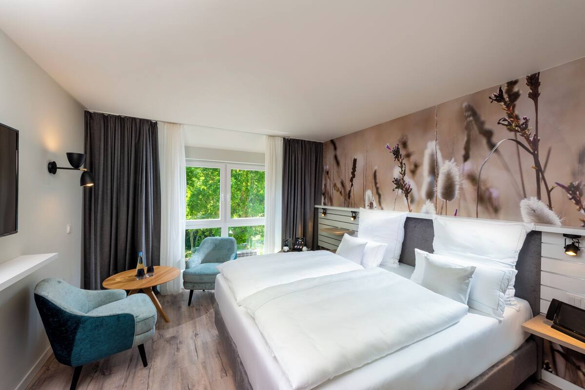 A modern hotel room with a large bed, a small desk, a chair, and a window with curtains.