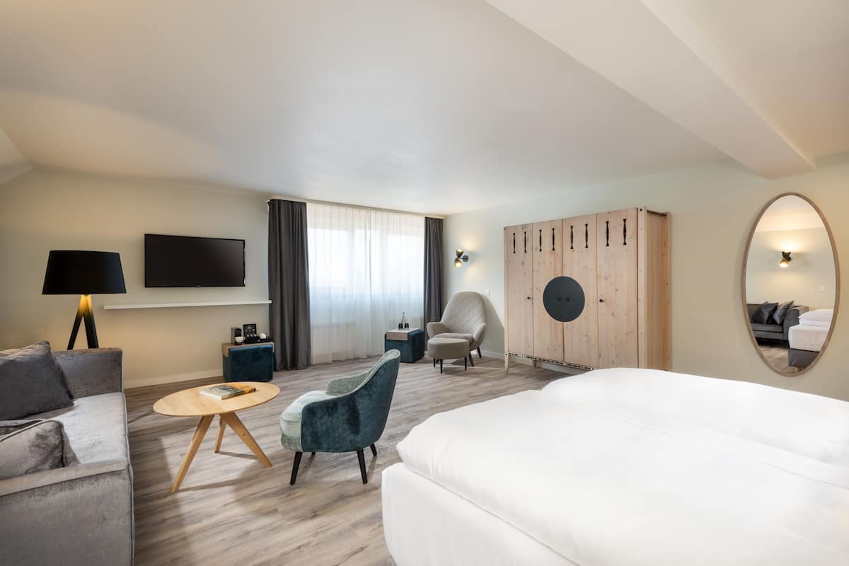 A modern hotel room with a large bed, a sofa, a round coffee table, a blue armchair, a TV mounted on the wall, a wardrobe, and a mirror on the wall.