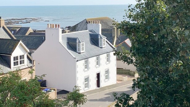 A white building with a gabled roof, a chimney, and a door, situated on a street with a view of the sea. - Beautiful Detached House, just meters from the sea (Gourdon)