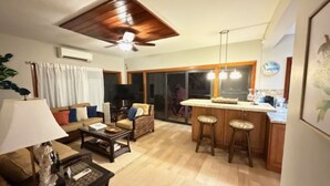 Ein Wohnzimmer mit Deckenventilator, Fernseher, Sofa und Couchtisch. Es gibt eine Küchenzeile mit einer Bar und Hockern.