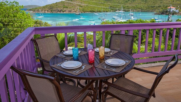 Ein runder Tisch mit vier Stühlen, einem violetten Geländer und Blick auf Boote auf türkisfarbenem Wasser.