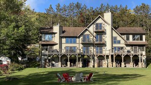 A large, multi-story building with a prominent balcony area, a grassy lawn, and outdoor seating. - Walk to Wisp! Flat Lakefront + Fire Pit and Huge Dock! (McHenry)