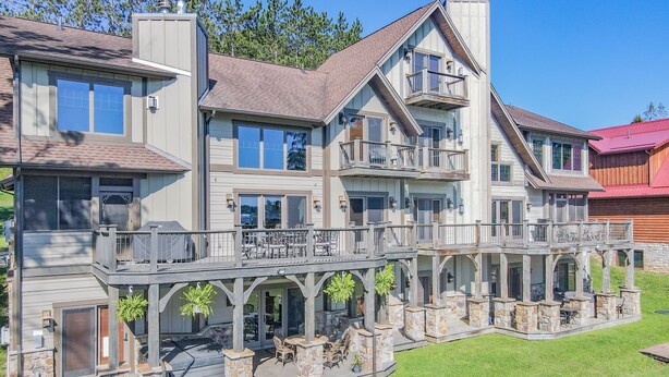 A large, multi-story house with a covered patio area, stone pillars, and outdoor furniture. - Walk to Wisp! Flat Lakefront + Fire Pit and Huge Dock! (McHenry)