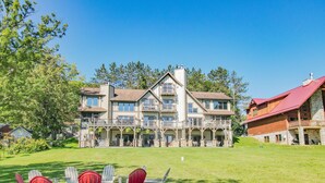 A large, multi-story building with a prominent stone facade, multiple balconies, and a well-maintained lawn with outdoor seating. - Walk to Wisp! Flat Lakefront + Fire Pit and Huge Dock! (McHenry)