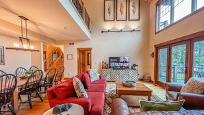 A spacious living room with a red sofa, a brown leather sofa, and a dining area with a wooden table and chairs. A television is mounted on the wall, and there are framed leaves on the wall above it. - Walk to Wisp! Flat Lakefront + Fire Pit and Huge Dock! (McHenry)