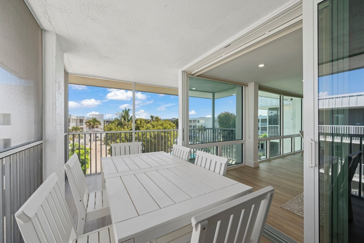 Un balcon avec une table et des chaises blanches, de grandes fenêtres et une vue sur des immeubles et des arbres.