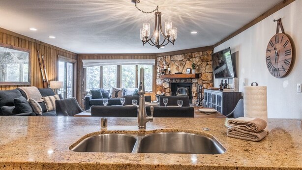 A spacious living area with a stone fireplace, a large wall clock, and a modern kitchen with granite countertops and stainless steel sinks. - Mountainback at Mammoth Lakes (Mammoth Lakes)