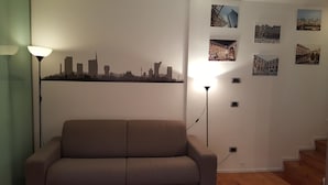 Un salon comprenant un canapé, une lampe et un mur décoré de photographies représentant des paysages urbains et des bâtiments.