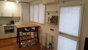 Une cuisine compacte avec des meubles blancs, un plan de travail en bois et un four encastré. On y trouve deux tabourets de bar noirs et une table en verre.
