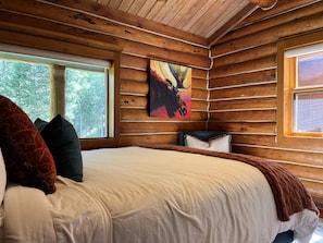 A log cabin bedroom with a large bed, two windows, and a painting of a moose. - Cozy, historic cabin in the woods, X-ski in/out, minutes to shops & restaurants (Big Sky)