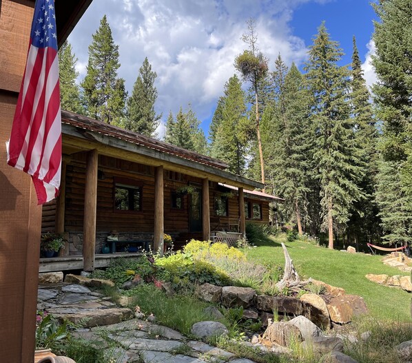 A log cabin with a porch, surrounded by a garden and tall trees. - Cozy, historic cabin in the woods, X-ski in/out, minutes to shops & restaurants (Big Sky)