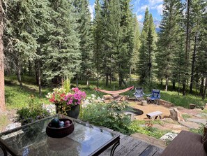A glass table with a bowl of fruit, a hammock, and two folding chairs in a forested area with a view of mountains. - Cozy, historic cabin in the woods, X-ski in/out, minutes to shops & restaurants (Big Sky)