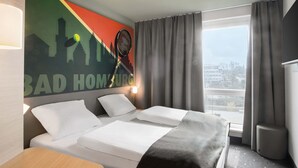 A hotel room with a bed, a desk, a window with curtains, and a wall with a cityscape mural.