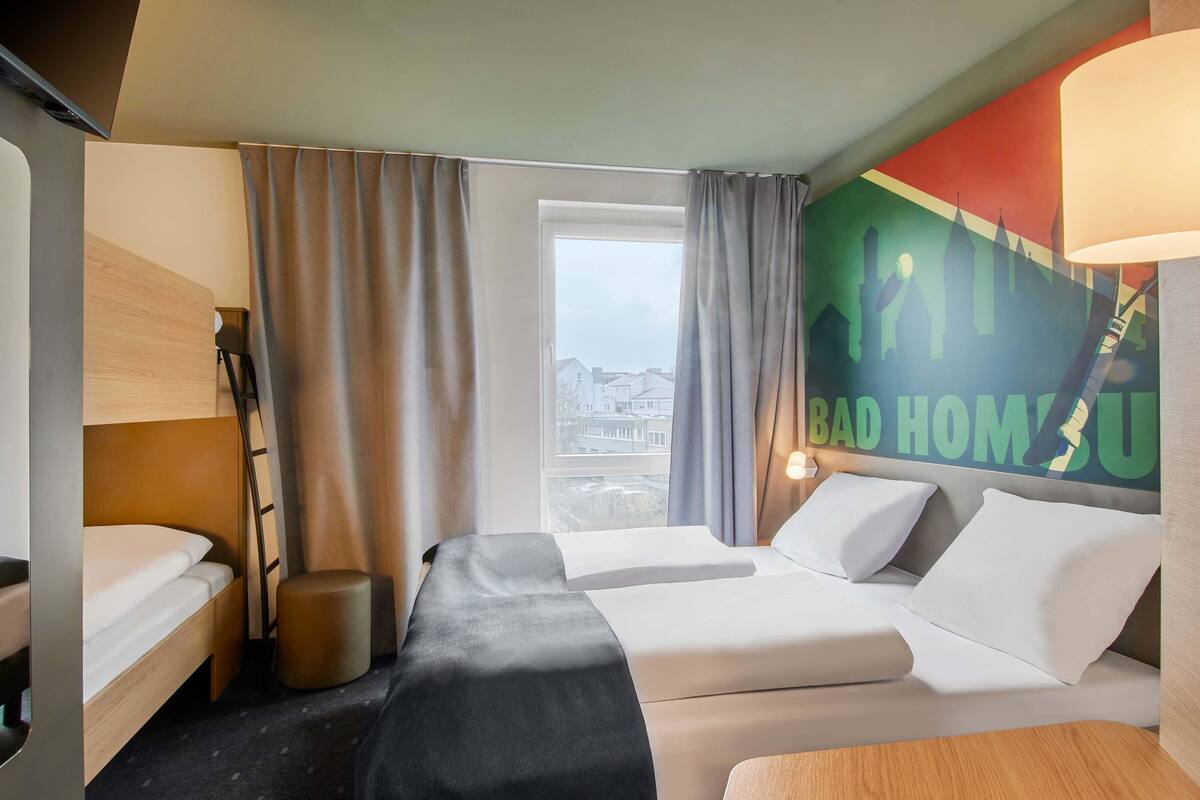 Une chambre d’hôtel avec deux lits, un bureau, une chaise, une fenêtre avec des rideaux et un mur orné d’une affiche représentant un paysage urbain.