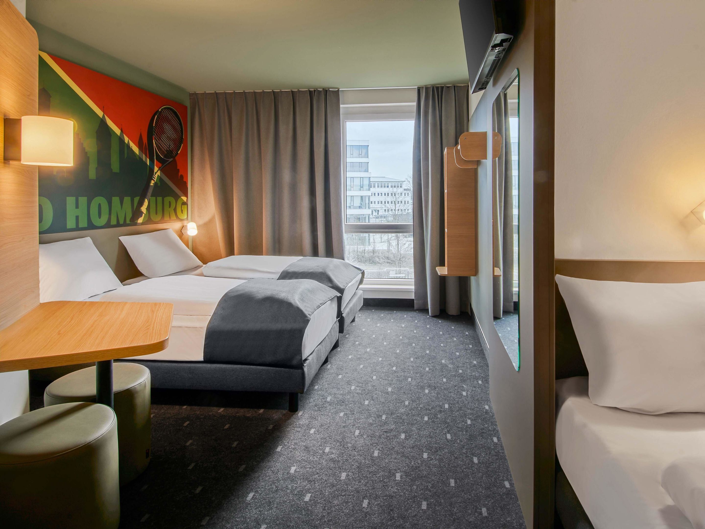 Ein Hotelzimmer mit zwei Betten, einem Schreibtisch, einem Stuhl und einem großen Fenster mit Blick auf die Stadt.