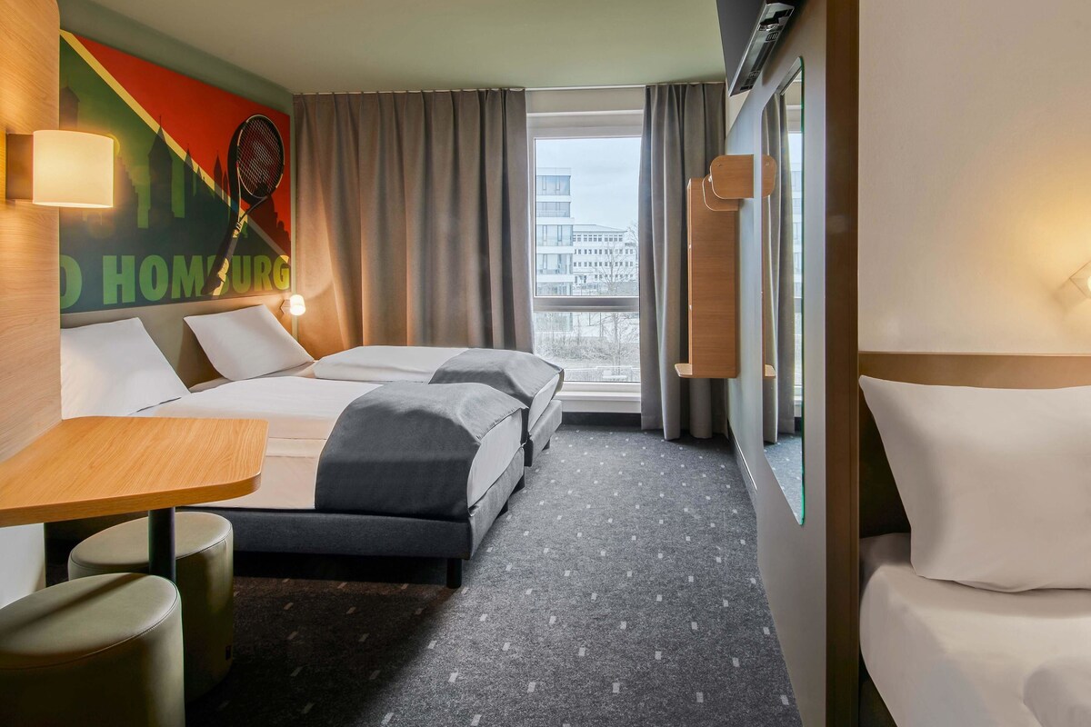 Ein Hotelzimmer mit zwei Betten, einem Schreibtisch, einem Stuhl und einem großen Fenster mit Blick auf die Stadt.